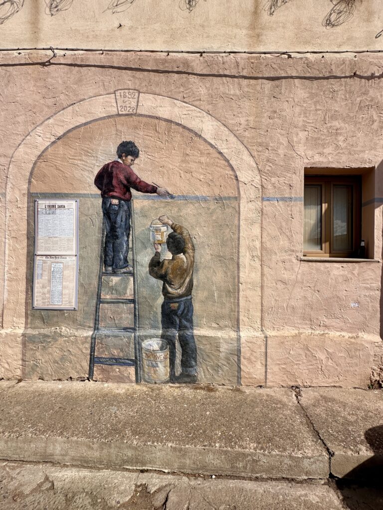 Murale a San Sperate con due ragazzi dentro un arco dipinto: uno sulla scala traccia una linea sul muro, l'altro regge un barattolo di colore, accanto a due pagine di giornale incorniciate