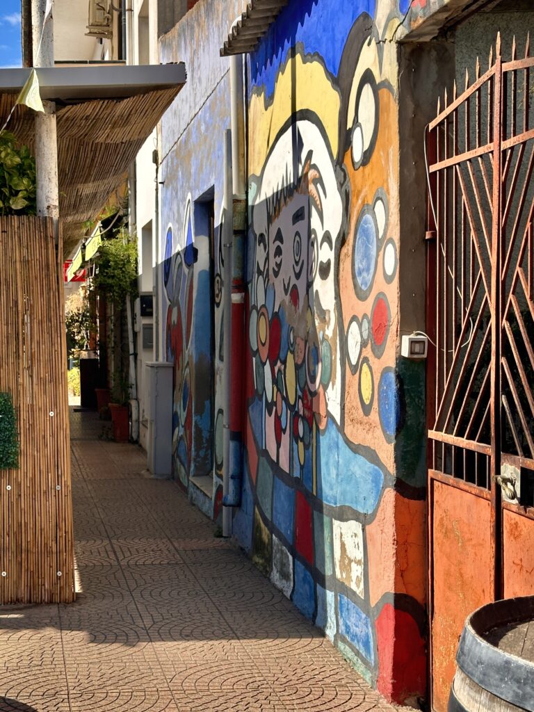 Murale colorato in un vicolo di San Sperate con un grande volto stilizzato circondato da cerchi rossi, blu e gialli, accanto a un cancello arrugginito e a una botte di legno