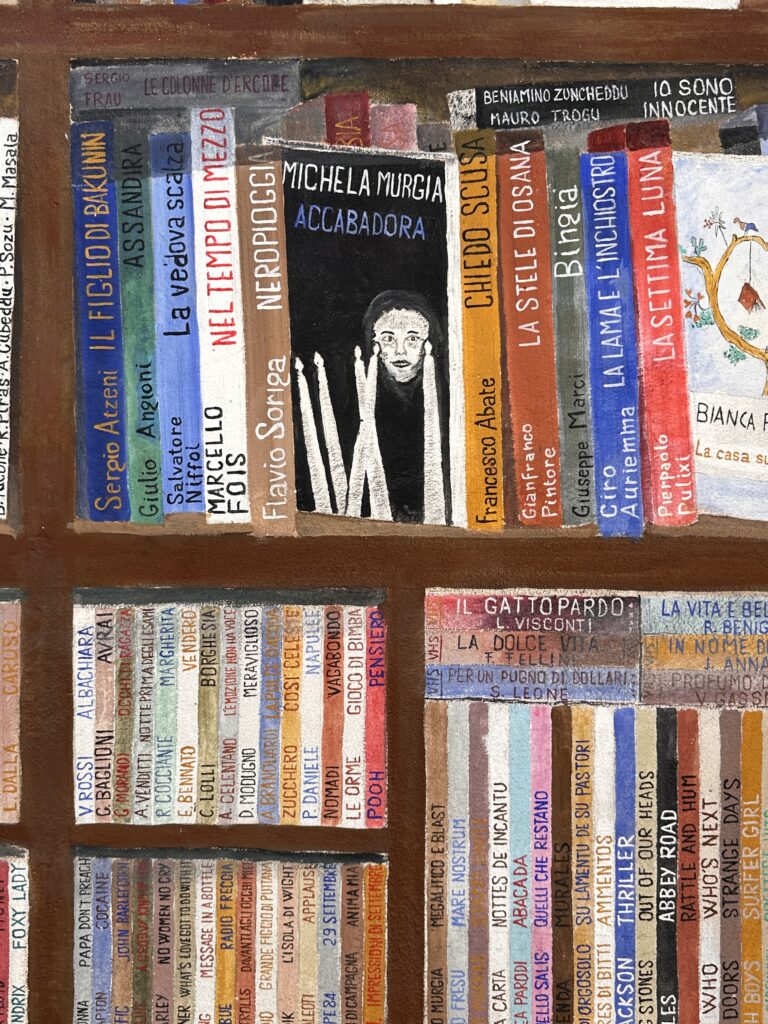 dettaglio del murale libreria a San Sperate sui dorsi della prima mensola c'è scritto "Cuncambias" con gli anni dal 2005 in poi. È il festival che le fonti legano all'omaggio a Sciola.