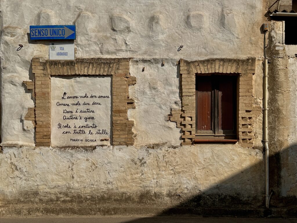 Poesia di Pinuccio Sciola scritta a mano in una finestra murata incorniciata di mattoni, su un muro bianco scrostato in via Arbarei a San Sperate