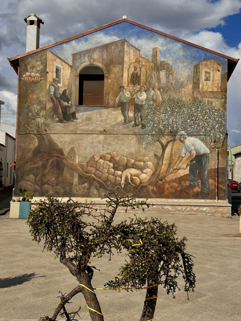 Murale dedicato al santo patrono di Angelo Pilloni in via Monastir, con processione, anziani seduti, frutteto e contadino che zappa