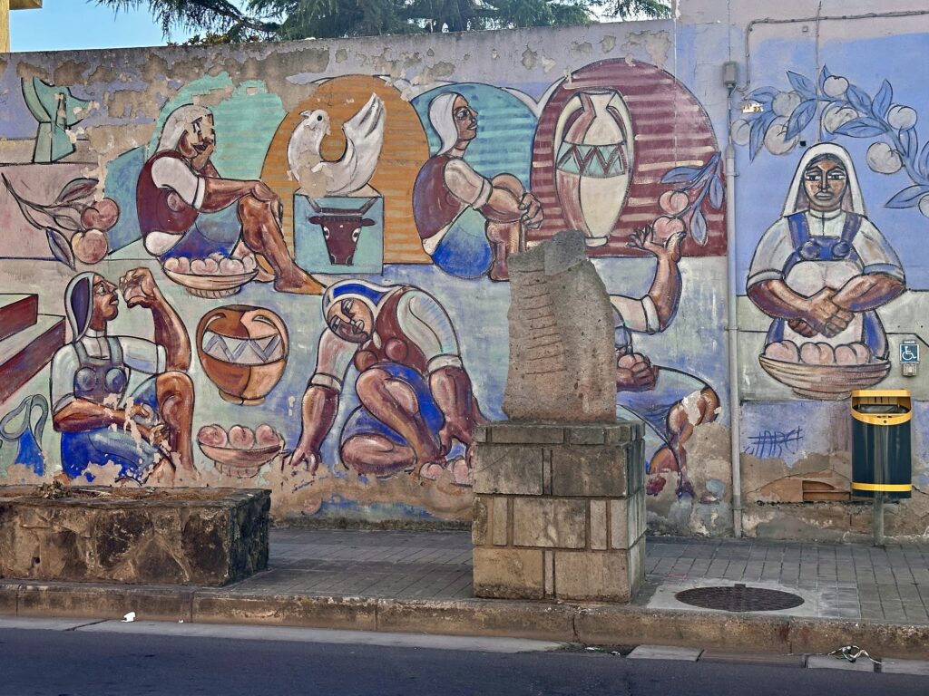 Murale con donne in abiti tradizionali sardi, ceste di pesche, anfore, una colomba e una testa di toro, con una scultura in pietra su basamento davanti al muro