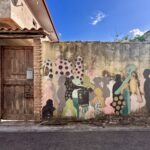 Murale con sagome umane colorate e motivi a pois accanto a un antico portone in legno, a San Sperate
