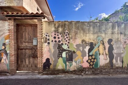Murale con sagome umane colorate e motivi a pois accanto a un antico portone in legno, a San Sperate