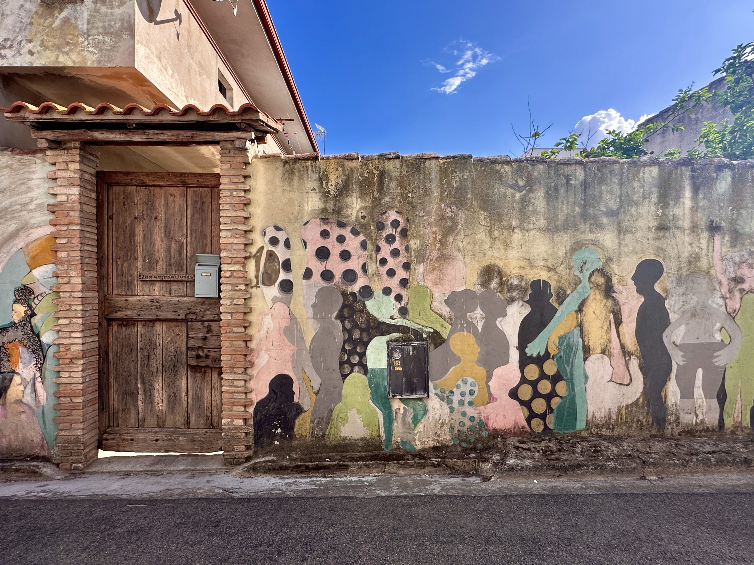 Murale con sagome umane colorate e motivi a pois accanto a un antico portone in legno, a San Sperate
