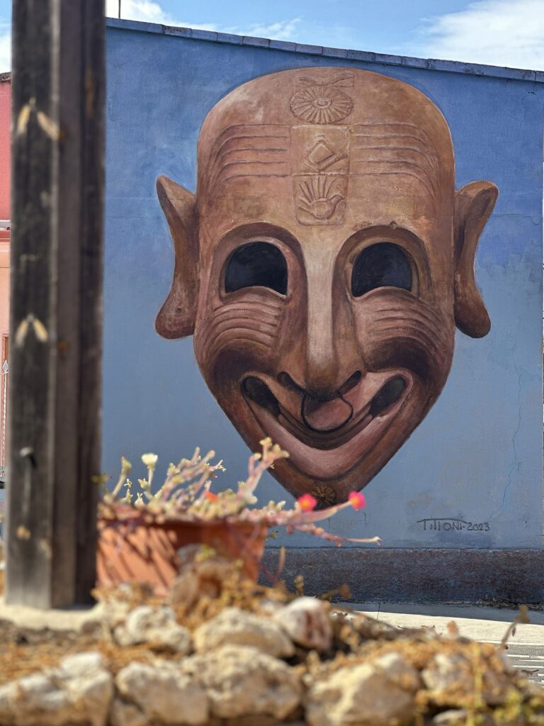 Murale della maschera ghignante firmato Pilloni 2023 su un muro blu a San Sperate, con sassi e fiori in primo piano