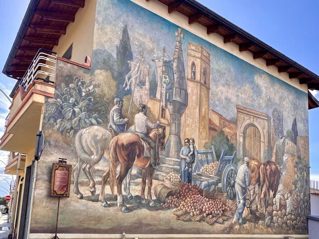 Murale Storia di San Sperate di Pinuccio Sciola in via Sassari, con processione a cavallo, stendardi, chiesa, carro di frutta e contadino con i buoi