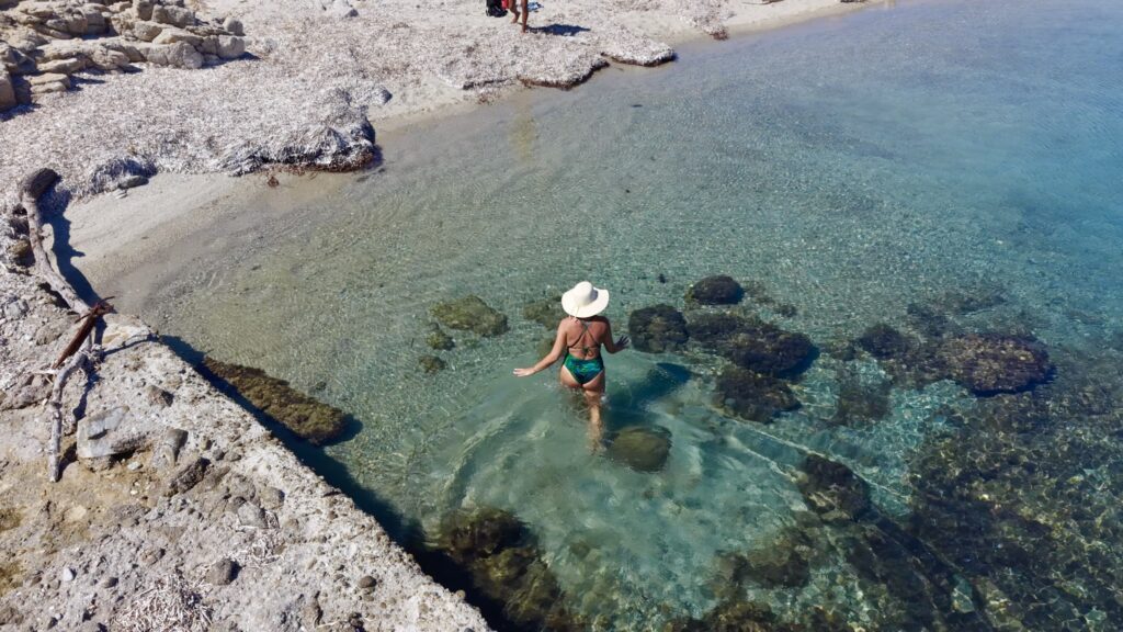 Donna con cappello che entra nell'acqua bassa e limpida accanto al pontile, tra rocce chiare e legni portati dal mare