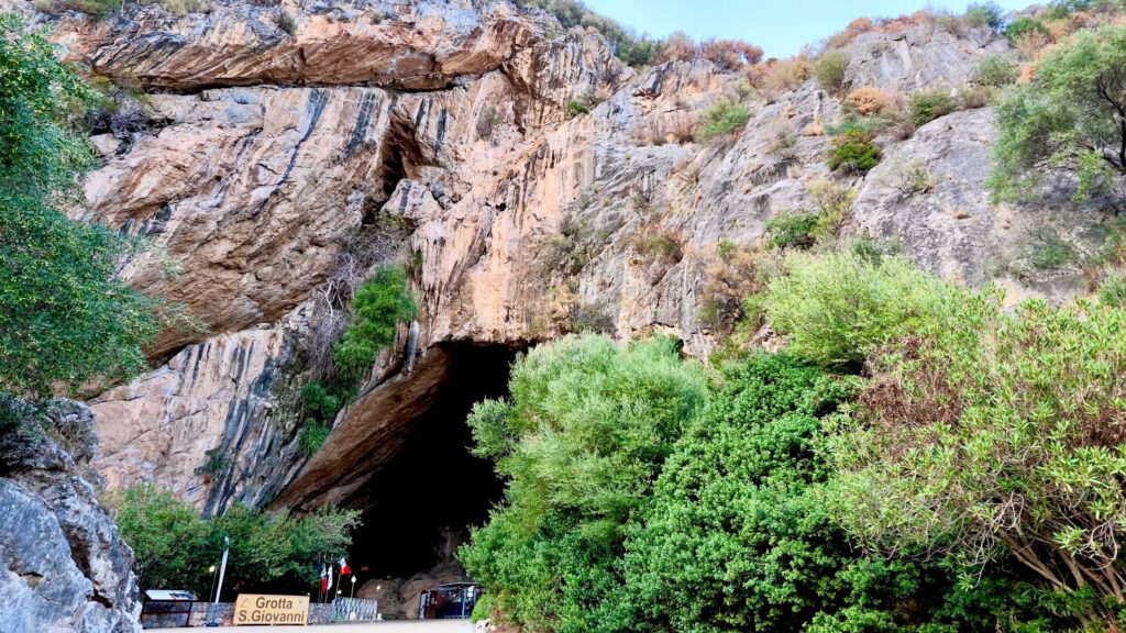 Cartello Grotta S. Giovanni e bandiere davanti all'ingresso sud, sotto un'alta parete di calcare con la macchia mediterranea