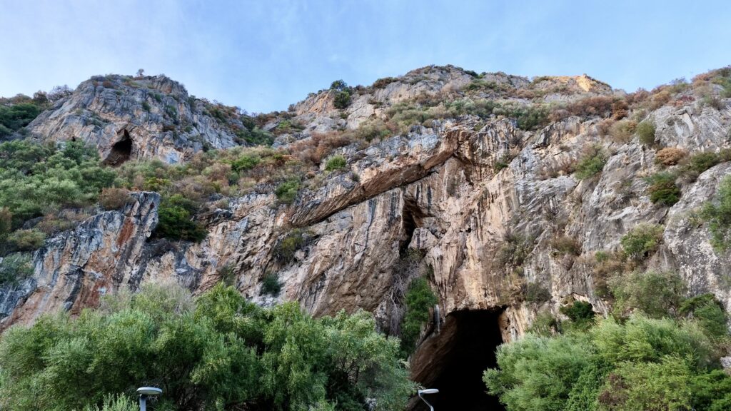 Ingresso sud della Grotta di San Giovanni a Domusnovas alla base di un'alta parete di calcare coperta di macchia mediterranea