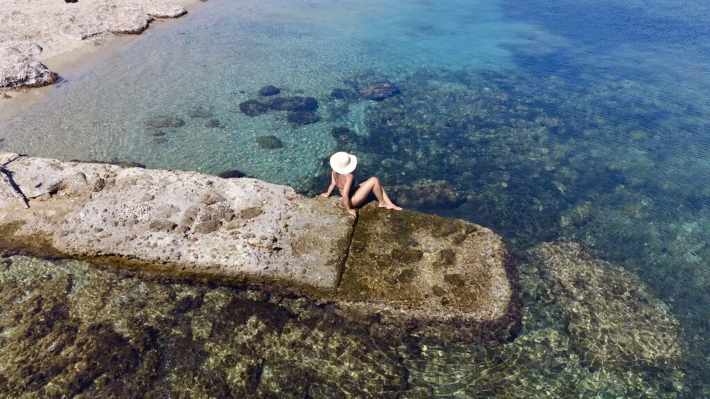 Donna in costume verde seduta sul bordo del pontile di pietra, con i riflessi del sole sull'acqua bassa