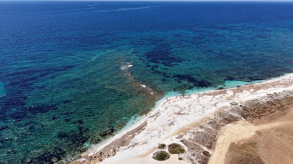 Vista dall’alto della spiaggia di Maimoni, con la riva chiara di quarzo, gli scogli bassi nell’acqua e il mare che sfuma dal turchese al blu