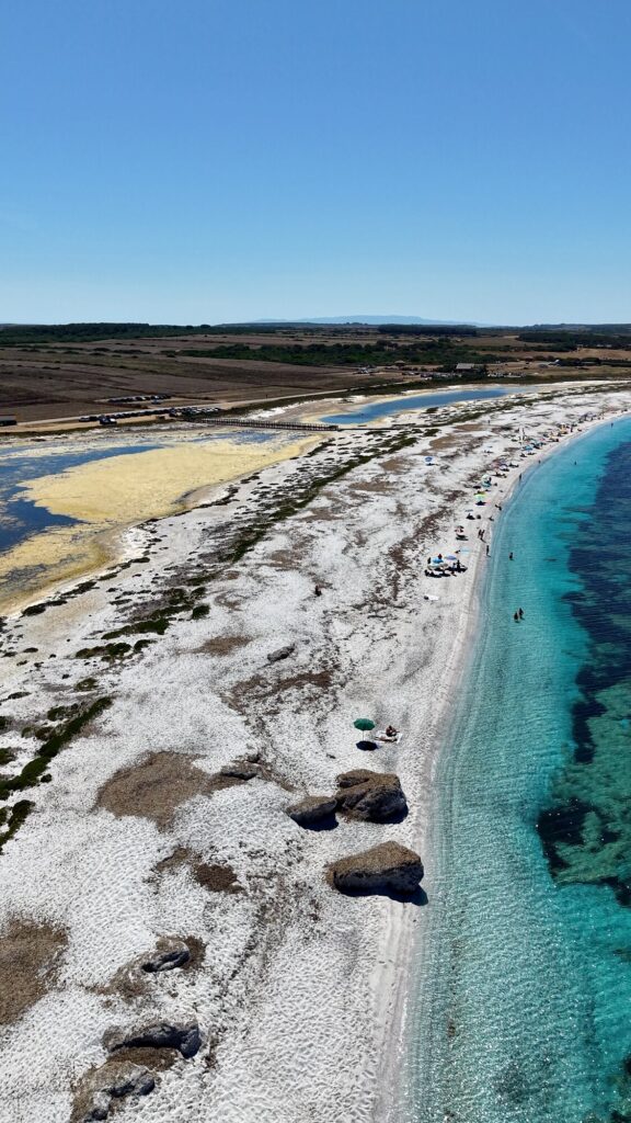 Vista dall’alto di Mari Ermi, con la spiaggia bianca di quarzo, gli scogli scuri sulla riva, il mare turchese e lo stagno alle spalle Didascalia: Mari Ermi è una lingua di quarzo bianco tra il mare e lo stagno.