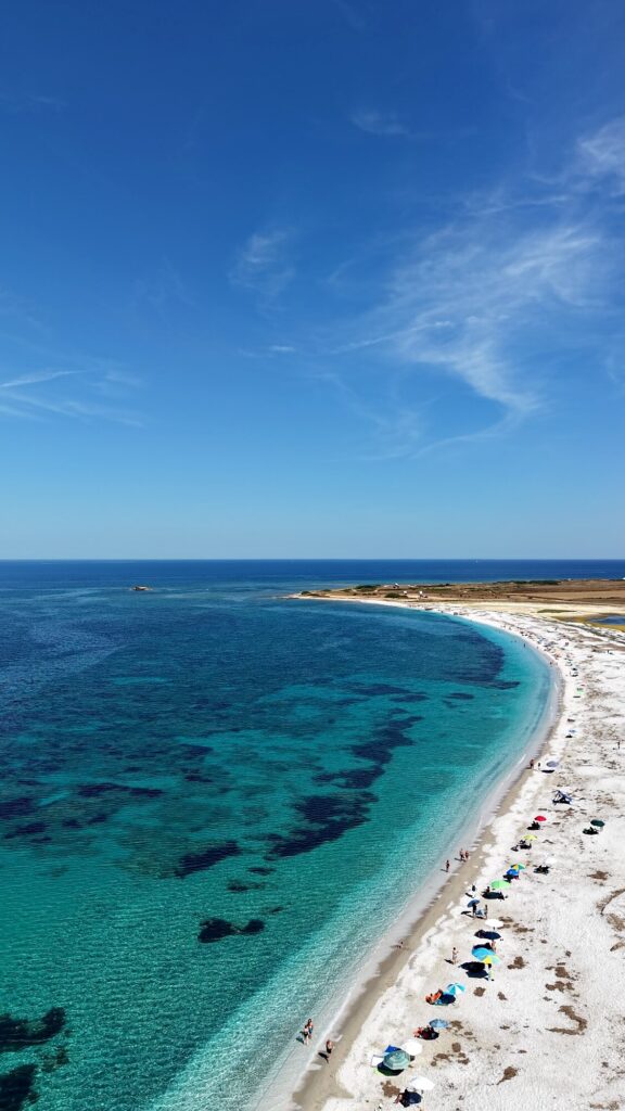 La lunga baia di quarzo vista dal drone, con il mare che sfuma dal turchese al blu, gli ombrelloni sulla riva e un cielo con nuvole leggere
