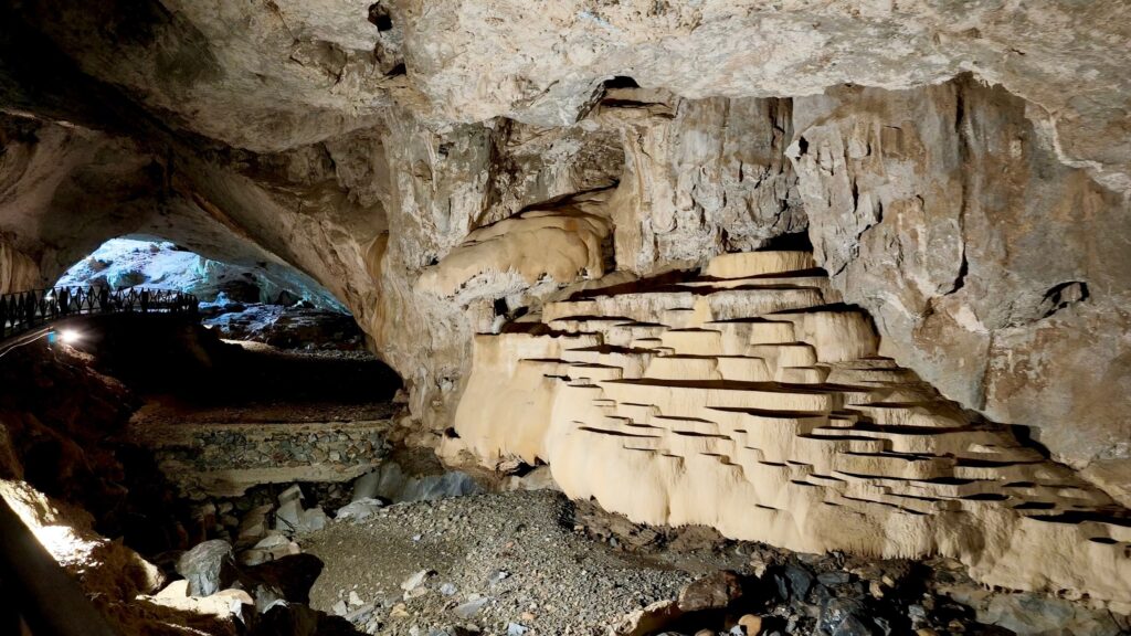 Grandi vaschette di calcare color crema accanto al letto del torrente, con la ringhiera e l'uscita nord della grotta sullo sfondo
